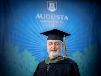 75-year-old Bill Nolan dress in his graduation regalia stands in front of a display of Augusta University
