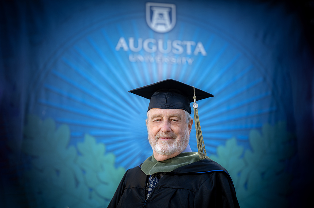 75-year-old Bill Nolan dress in his graduation regalia stands in front of a display of Augusta University