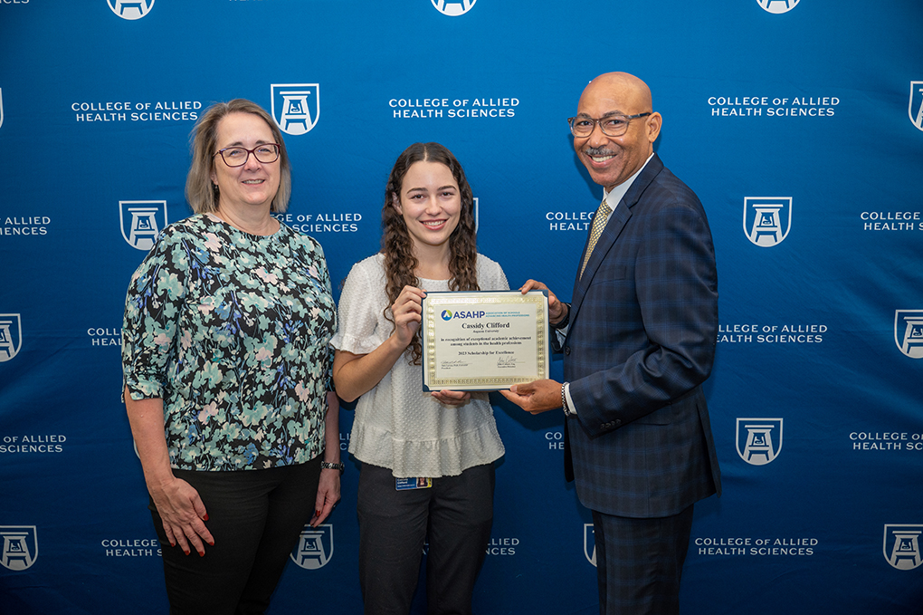 a woman wearing a flowered blouse, dark pants and glasses and a man in a black and dark blue checkered suit and glasses present an award to a young woman in a white blouse and dark pants in front of a dark blue backdrop with white AU and College of Allied Health Sciences logos