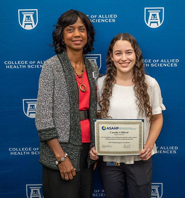 a woman wearing a belted red blouse and dark gray and light gray cardigan stands with a young woman in a white blouse and dark pants holding her award in front of a dark blue backdrop with white Augusta University and College of Allied Health Sciences logos
