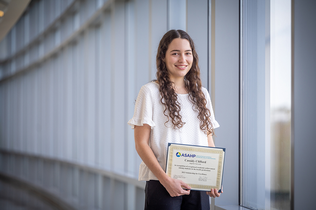 a woman in a white blouse and dark pants with long curly hair holds the Scholarship of Excellence award she received while standing beside a row of windows