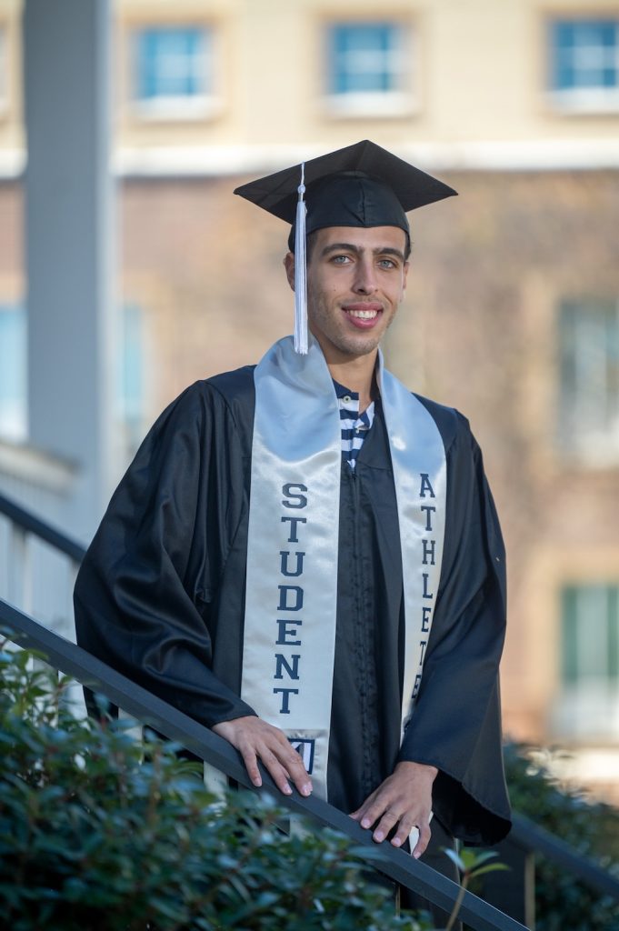 Graduate standing on steps wearing his graduation regalia including a gray student-athlete stoll with the AU logo on it