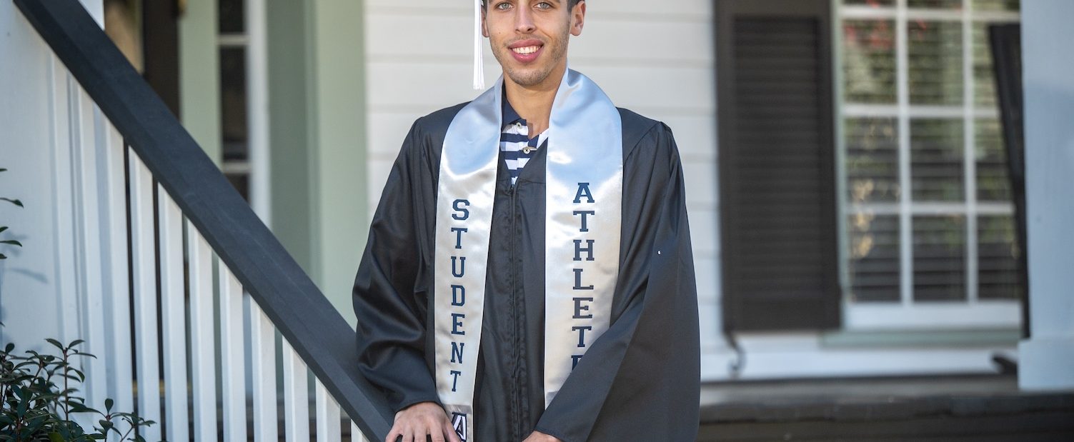 Graduate in his communication graduation regalia and student-athlete stoll poses on steps