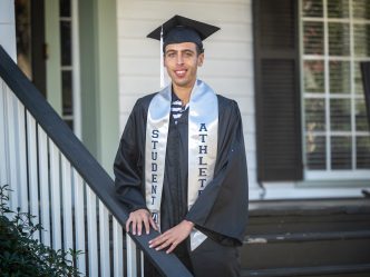 Graduate in his communication graduation regalia and student-athlete stoll poses on steps