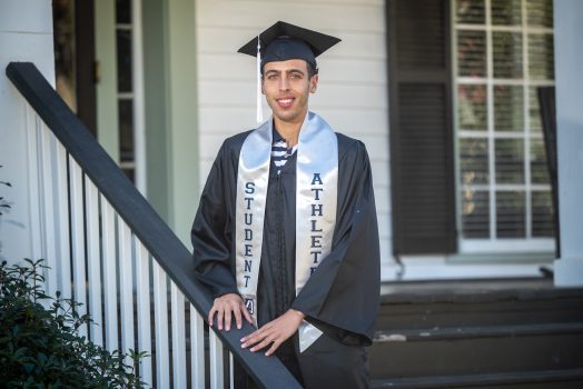 Graduate in his communication graduation regalia and student-athlete stoll poses on steps