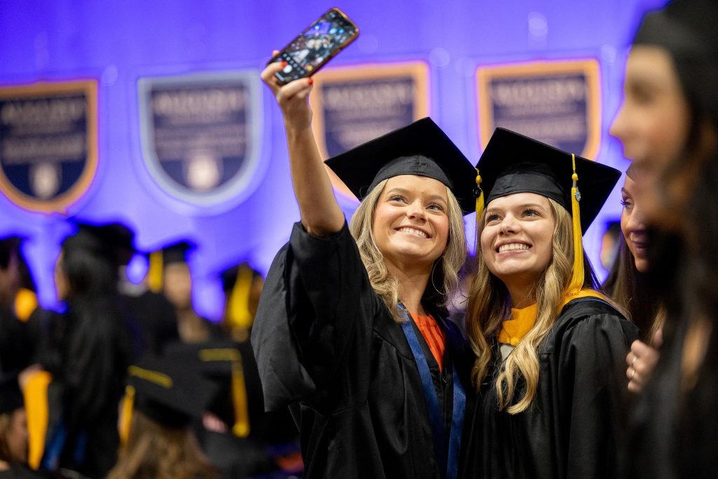 Two young women wearing graduation caps and gowns smile as one holds her cell phone up to take a photo.