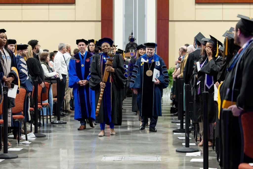 College professors wearing commencement garments of hats and robes walk down an aisle with a woman holding a wooden torch at the front.