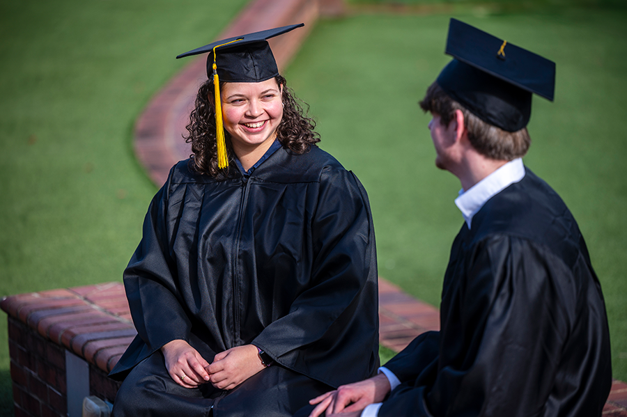 A woman in a graduation cap and gown sits on a low brick wall talking with a man, also in a graduation cap and gown.