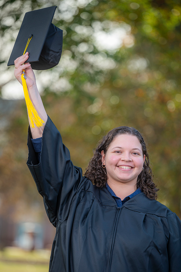 A woman wearing a graduation gown holds up a graduation cap above her head while smiling.