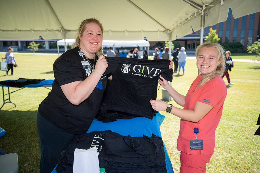 A woman hands another woman a short-sleeve t-shirt that reads IGIVE with the Augusta University logo on it.