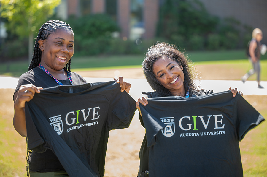 Two women hold up short-sleeve t-shirts that reads IGIVE with the Augusta University logo on it.