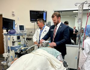 A man in a suit stands at the head of a gurney holding an anesthesia mask to a dummy patient while a woman in a lab coat watches a screen to his left