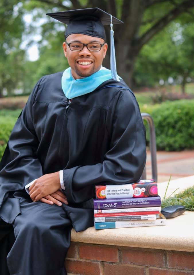 A male college student wears a graduation cap and gown while seated next to a large stack of textbooks. 