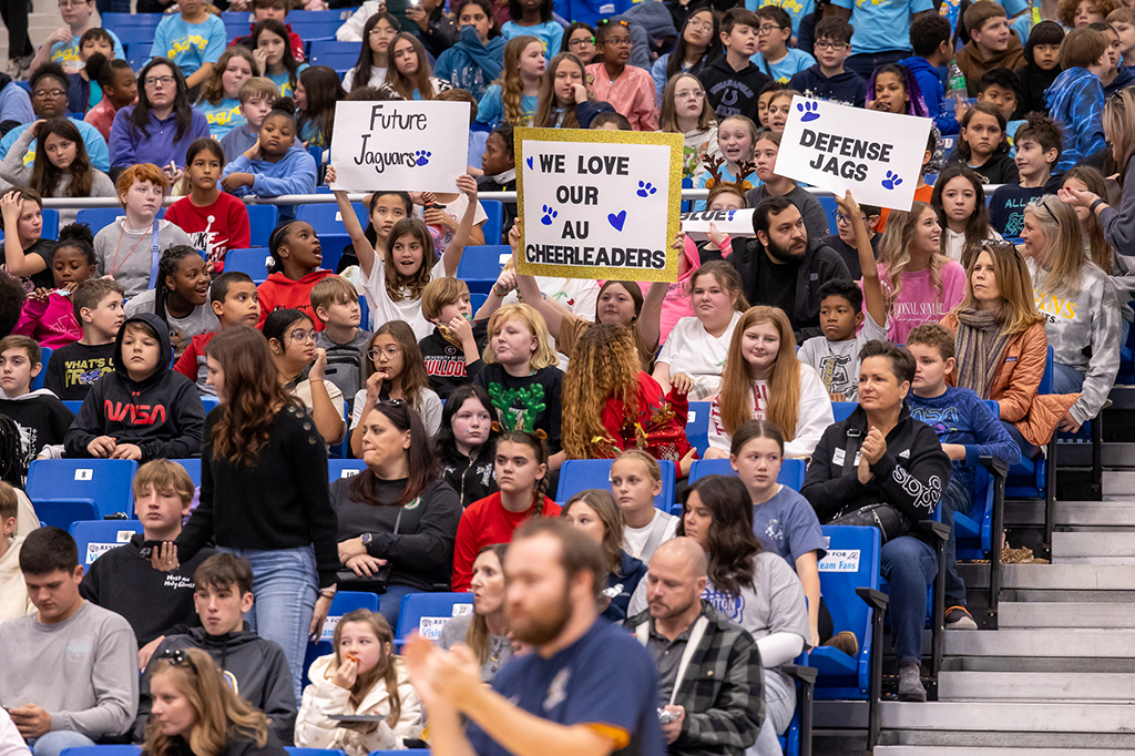 Children sitting in stands at an indoor basketball game while holding signs to support Augusta University's men's basketball team.