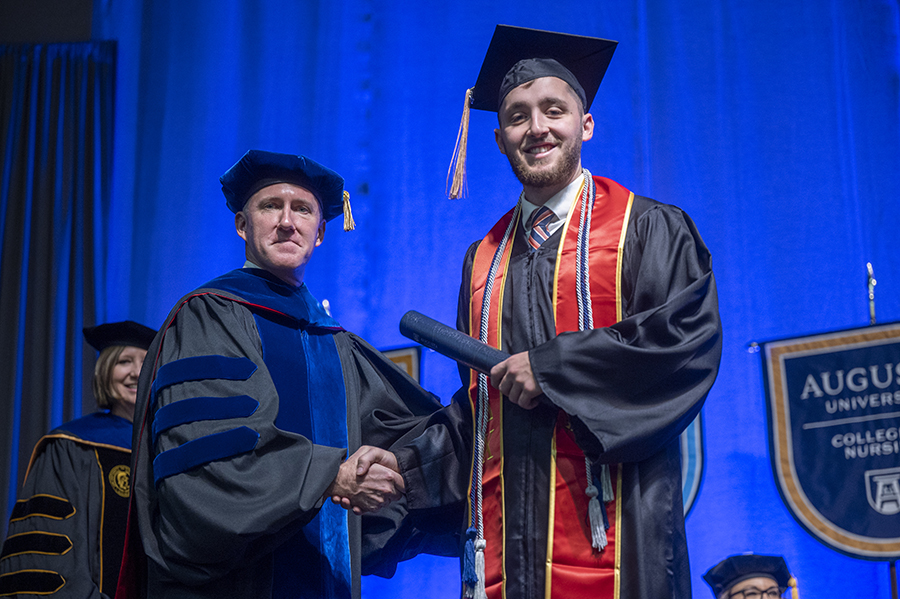 Two men wearing graduation caps and gowns shake hands while posing for a photo.
