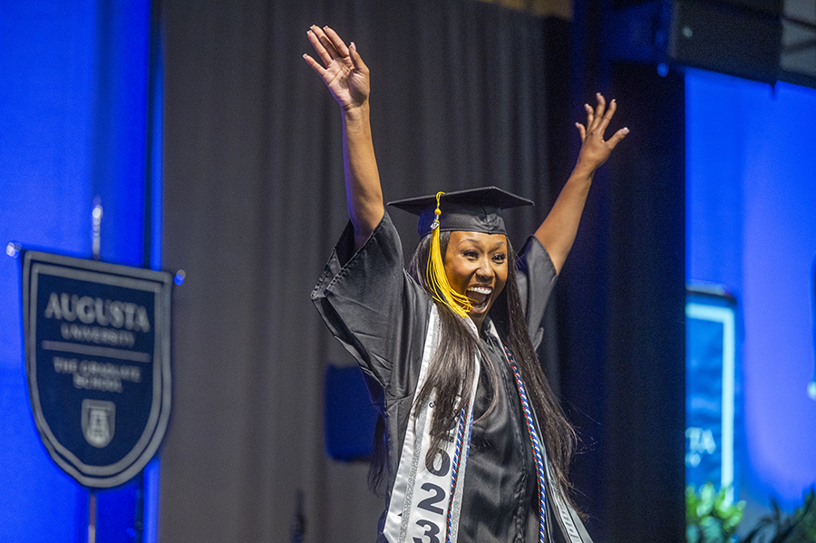 A young woman holds her hands up in celebration as she crosses a stage to accept her college diploman.