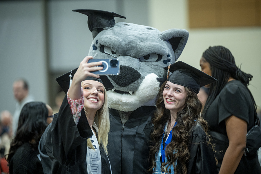Two young women pose with a college mascot as one of the women holds up her cellphone to take a selfie.