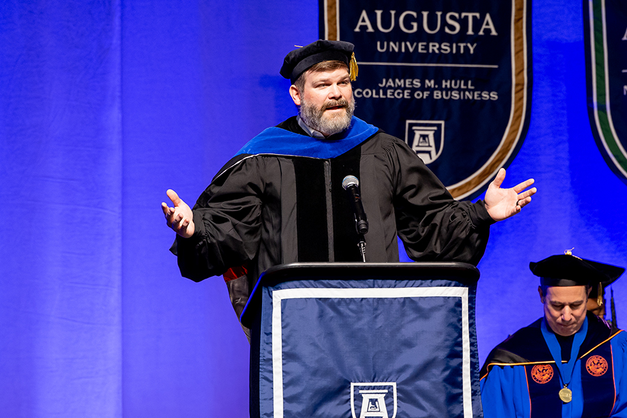 A man wearing a graduation cap and gown stands at a podium and delivers a speech while holding his hands out wide.