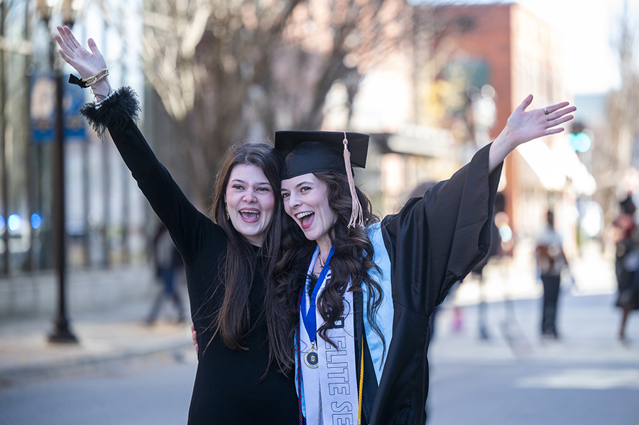 Two women, one wearing a graduation cap and gown, celebrate outside.