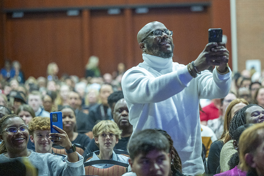A man stands up among a large seated crowd to take a photo with his cellphone. 