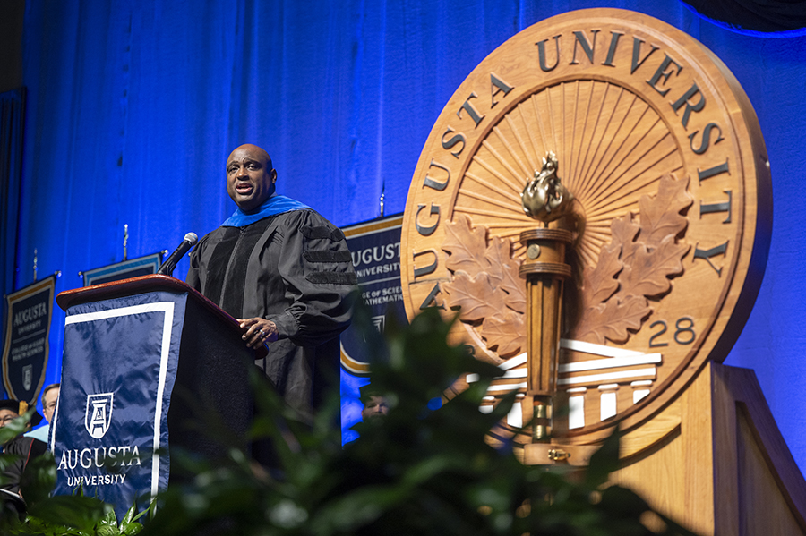 A man wearing a graduation gown stands at a podium to give a speech.
