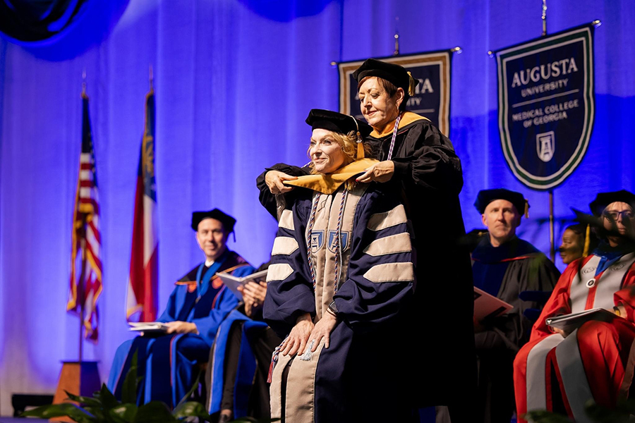 A woman bends at the knees to allow another woman to place a colorful garment around her neck signifying she has completed her Doctor of Nursing Practice degree.