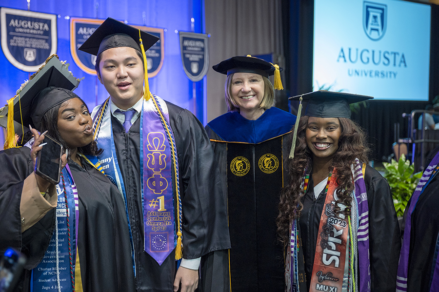 Three women and one man pose for a group photo. All four are wearing graduation caps and gowns.
