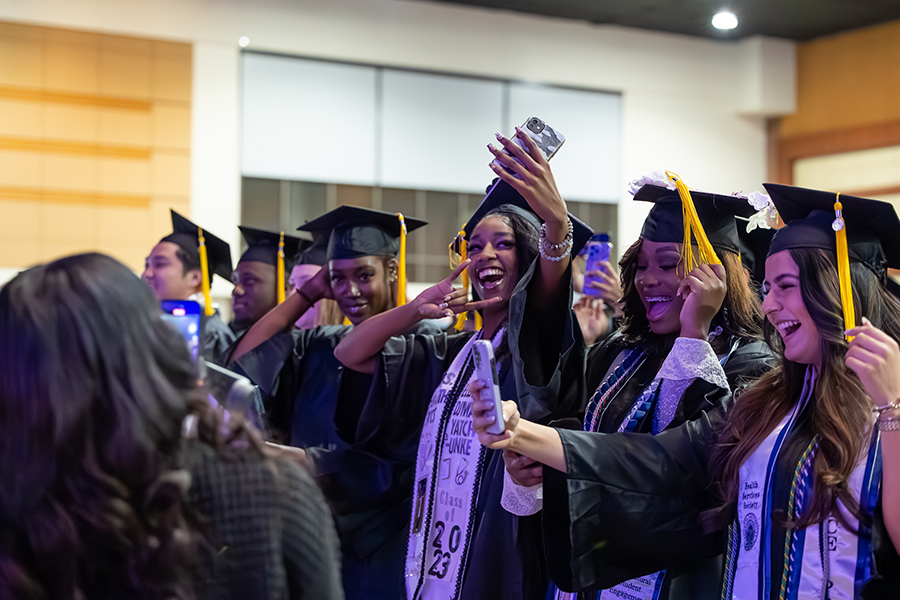 Four young women celebrate having graduated from college while wearing their caps and gowns. Two women hold up their cellphones for a selfie, while another moves her tassel from right to left.