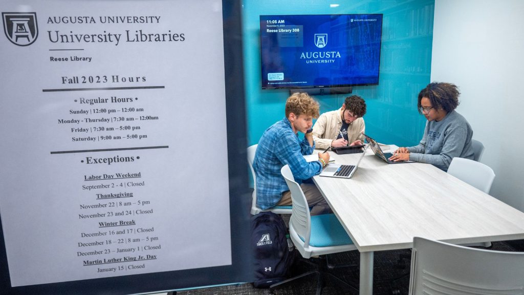 Three students work at a desk inside one of the new study rooms with a sign for AU Libraries, including hours of operation, in the foreground.