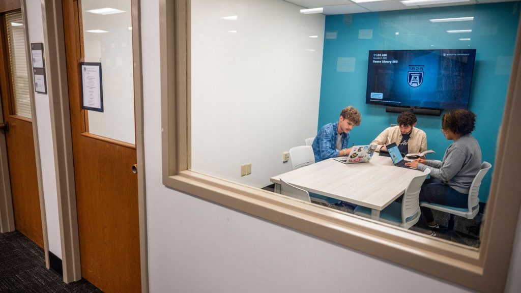 Three students sit working at a table inside one of the new study rooms with a large monitor on the wall behind them