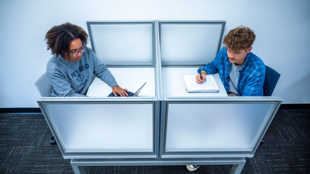 Two students work at  desk facing each other