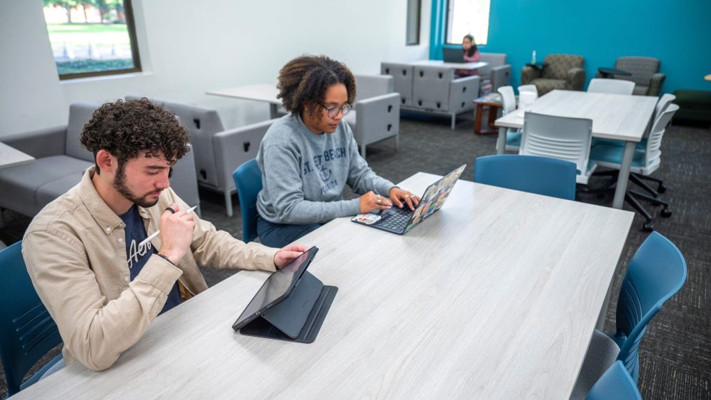 Two students sit at a table working, one on a laptop and the other on a tablet.