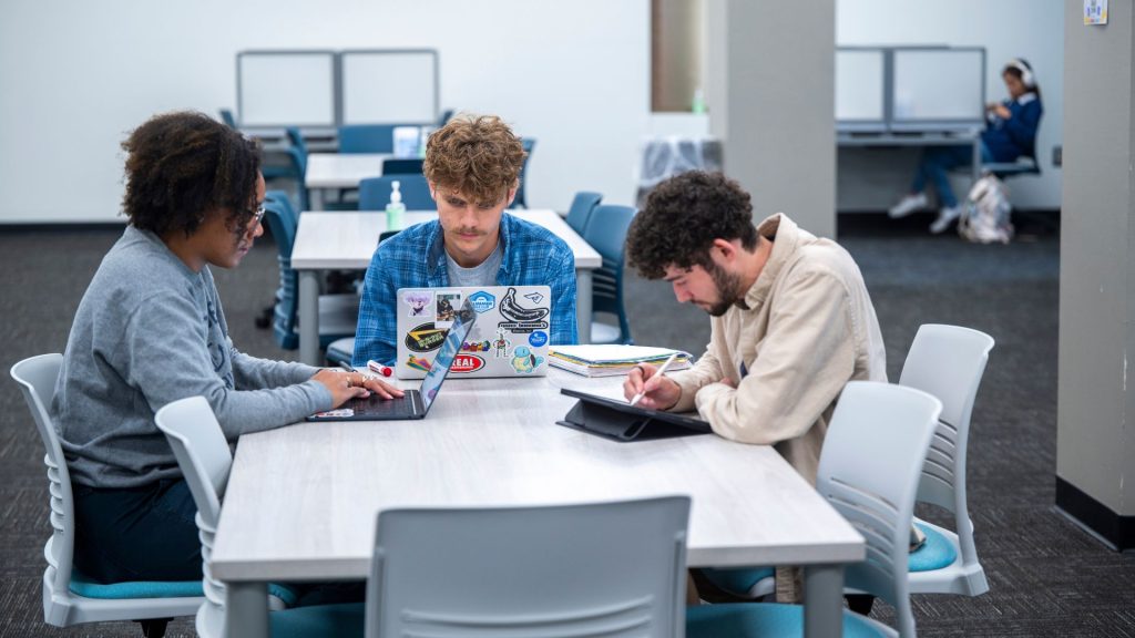 Three students sit at a table working on laptops and tablets.
