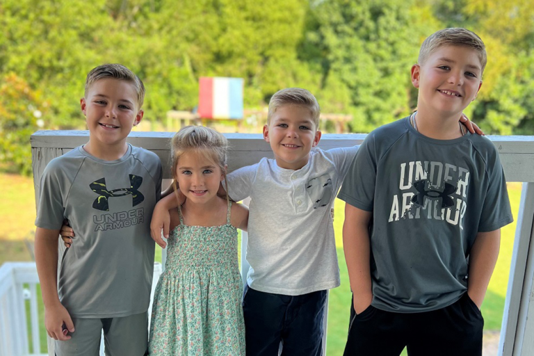 Four kids stand on a porch with their arms around each other and smile for a photo.