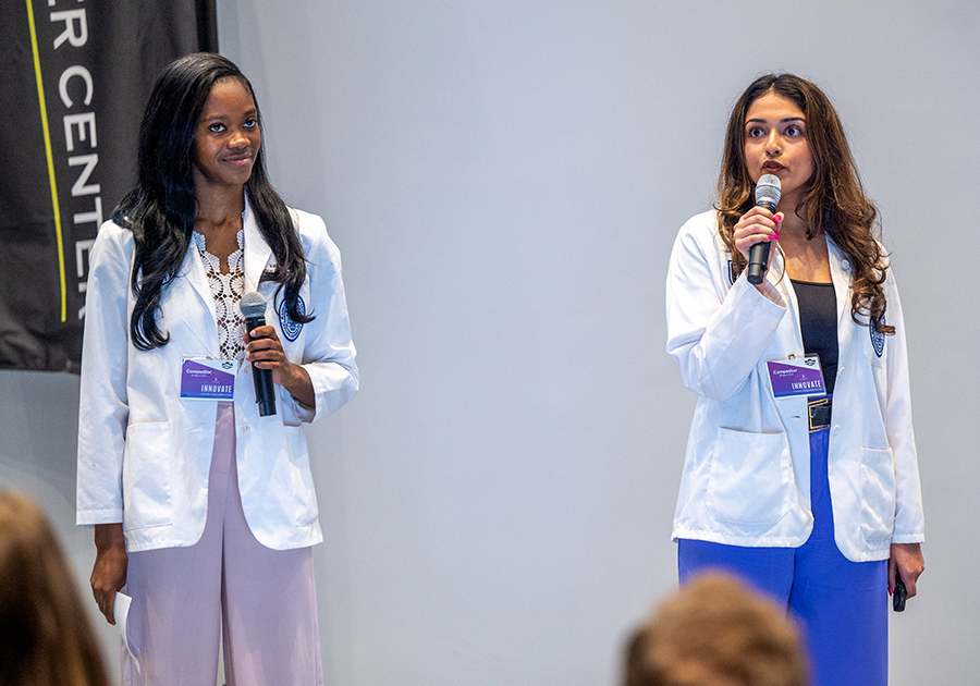Two medical students wearing their white coats hold microphones while presenting in front of an auditorium.