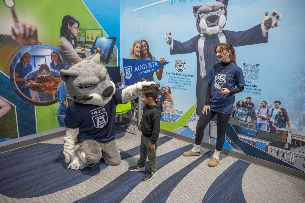 A college mascot depicting a jaguar interacts with a young child while a college student looks on and laughs.