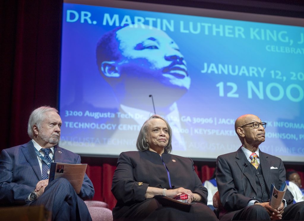 Two men and a woman sit on a stage during a Dr. Martin Luther King Jr. celebration