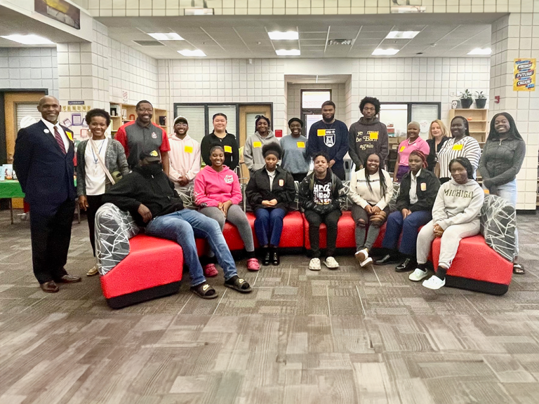 A group of students and teachers and administrators pose for a photo on and around a couch inside a school library