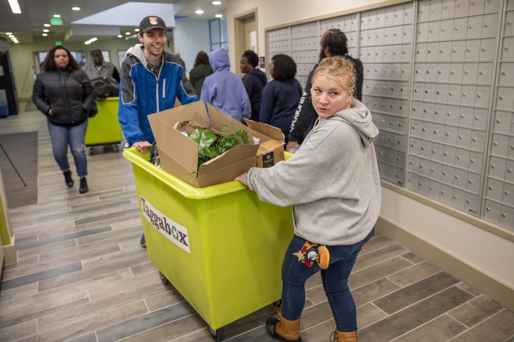 A young man and woman are inside pushing a bin filled with dorm room material
