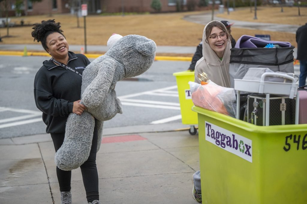 Two smiling young women move a bin and a big stuffed animal into a dorm