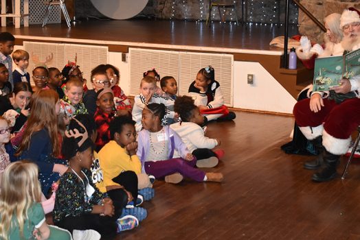 Santa Claus sits on the right reading a book to a large group of children seated on the floor in front of him