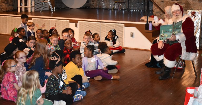 Santa Claus sits on the right reading a book to a large group of children seated on the floor in front of him