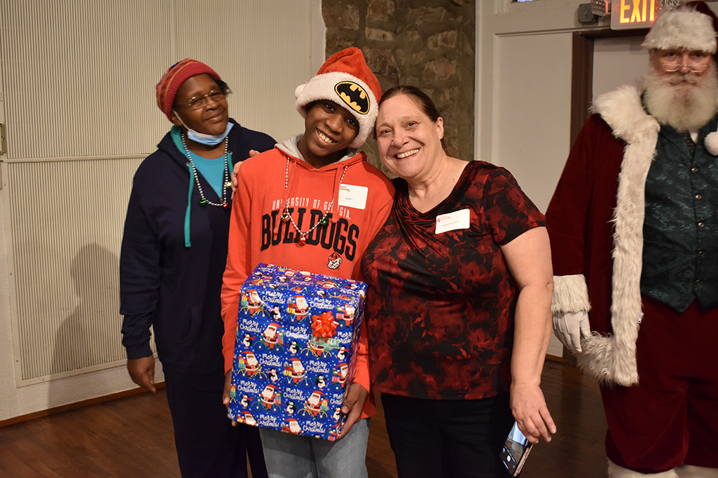 a boy with a Batman Santa hat and Bulldogs sweatshirt holds a present and smiles next to a woman in a red and black shirt as his grandma and Santa Claus look on