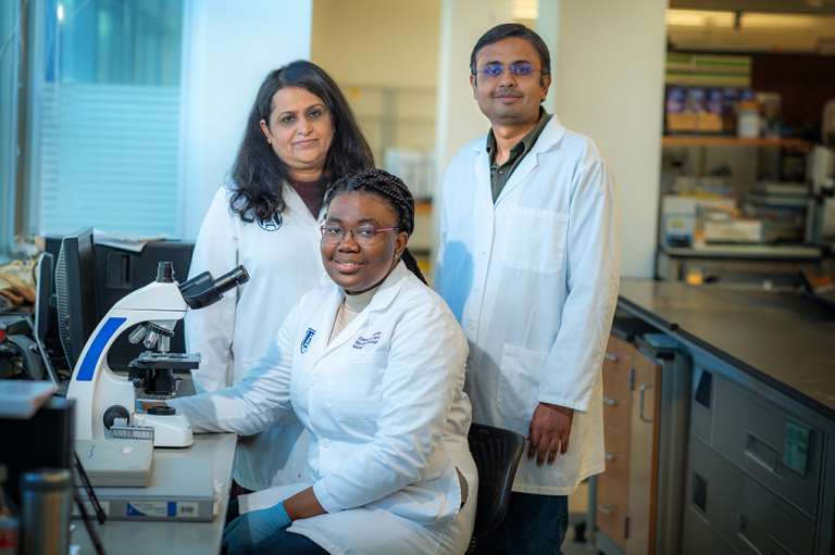 Two women and one man in scientific lab coats pose for a photo in their lab with a microscope nearby.