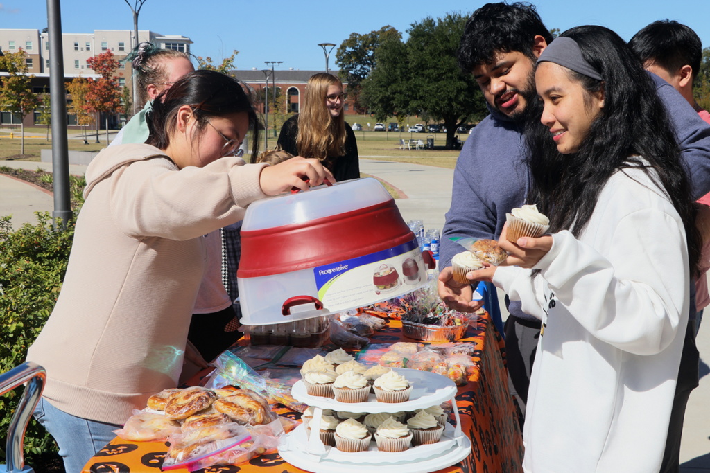 multiple students surround a table outside with an orange and black pumpkin tablecloth filled with various baked goods. The woman in the front on the right wearing a long white sweatshirt holds a cupcake
