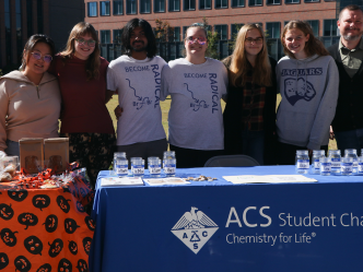 A professor stands with his students behind a club table for the American Chemical Society