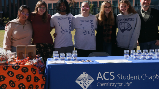 A professor stands with his students behind a club table for the American Chemical Society