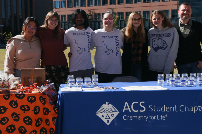 A professor stands with his students behind a club table for the American Chemical Society