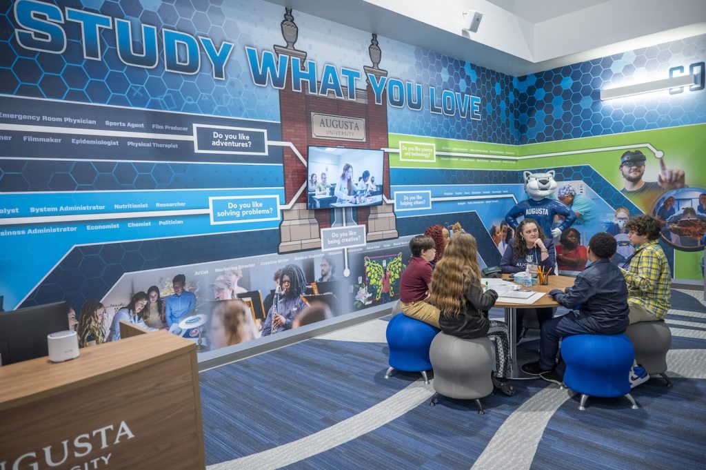 Four middle school students sit on chairs while two adult women listen to their discussion. On the wall behind them is the Study What You Love pathway that the kids can use to find careers that may fit their interests.
