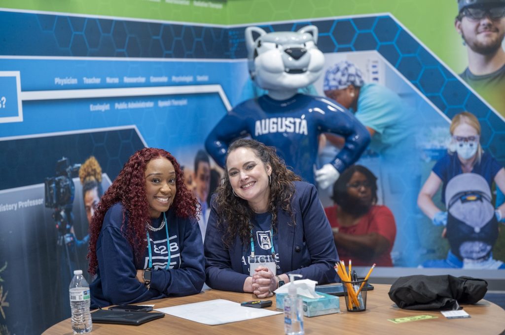 Two women sit at a table and smile for a photo with a statue of the Augusta University mascot Augustus standing behind them.